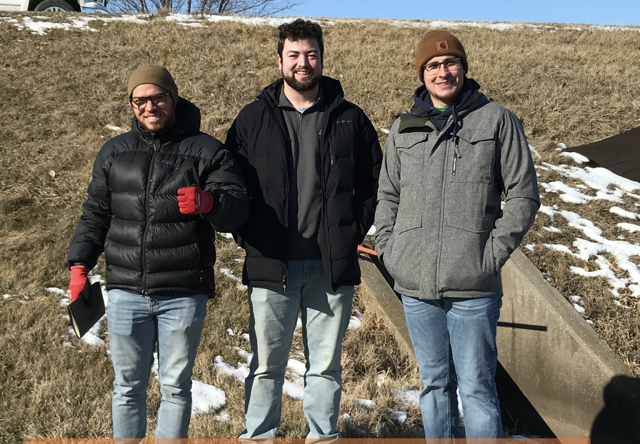 Three young people wearing winter coats and hats stand outside with a small patch of snow and bare ground behind them. 