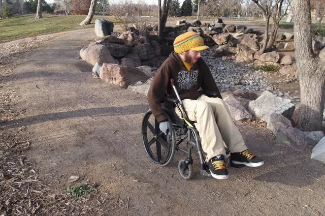 A young man in a yellow hat, dark sweatshirt, and tan pants in a wheelchair on an outdoor dirt path.