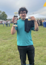 Young man with dark hair holding a trophy in a green field.